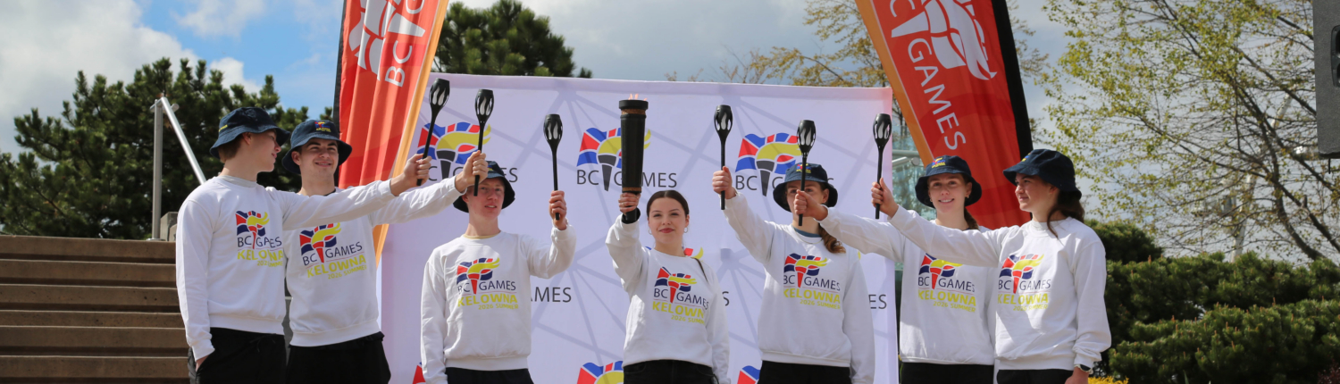 Seven young athletes in BC Games Kelowna 2026 sweatshirts raise torches together at the torchlighting ceremony at Jim Stuart Park, with BC Games banners in the background.