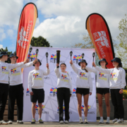 Seven young athletes in BC Games Kelowna 2026 sweatshirts raise torches together at the torchlighting ceremony at Jim Stuart Park, with BC Games banners in the background.